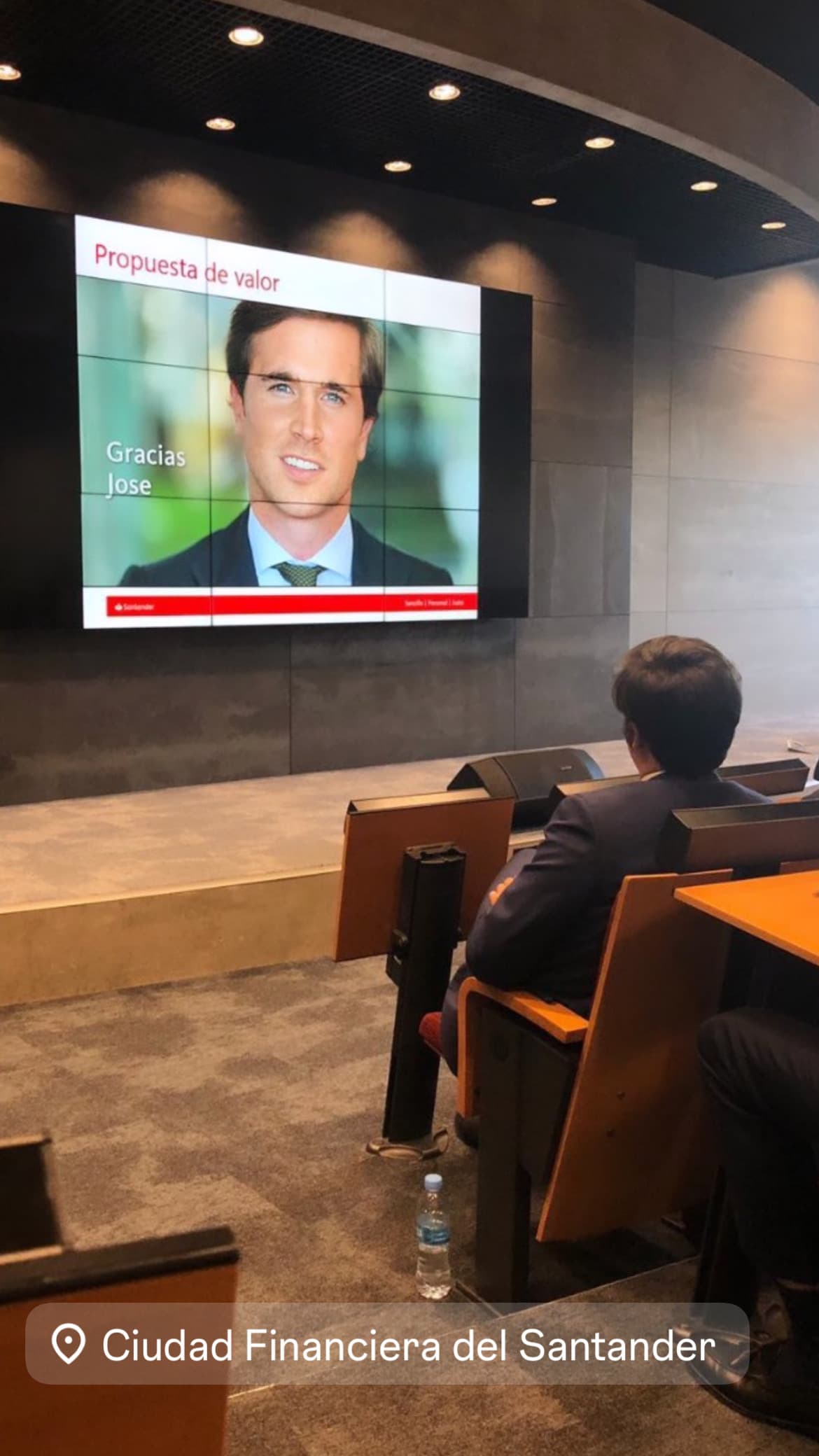 Hombre sentado en un auditorio observando una pantalla con un retrato y texto corporativo.