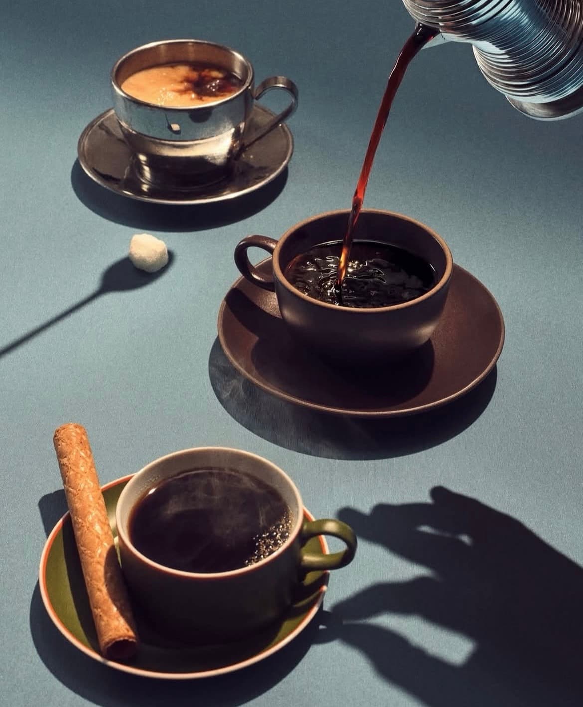 Café vertiéndose en una taza oscura junto a otras tazas, un barquillo y azúcar.