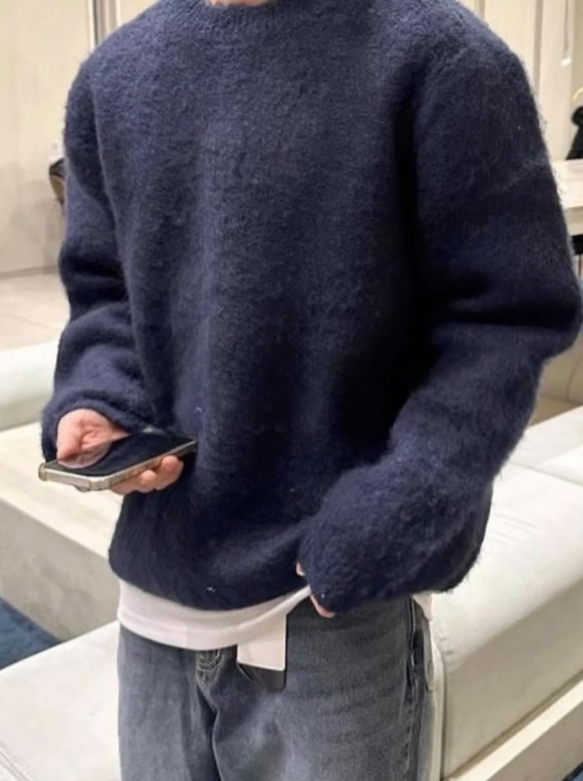 Persona con suéter azul marino de lana, jeans grises y sosteniendo un teléfono inteligente.