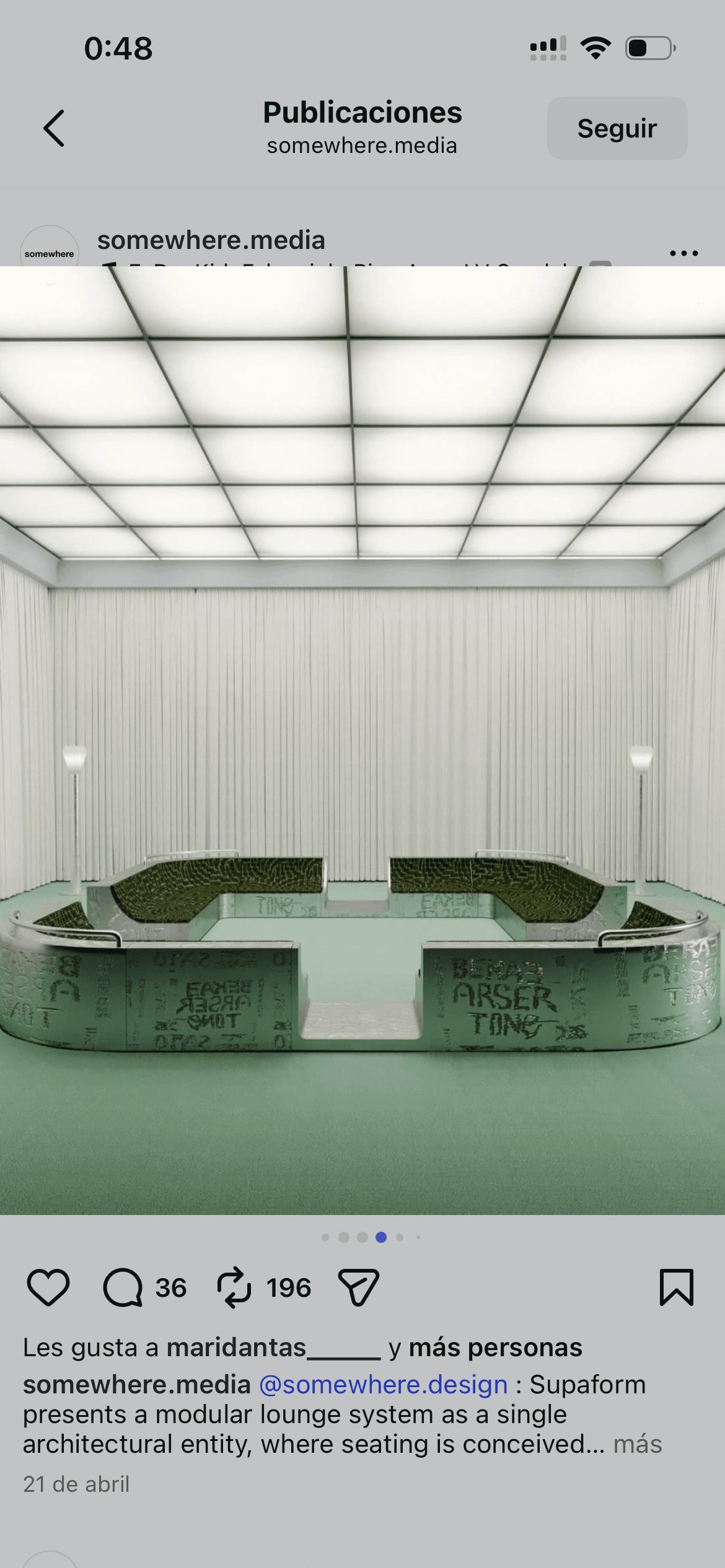 Sofá modular metálico con cojines verdes en sala blanca minimalista con techo cuadriculado iluminado.