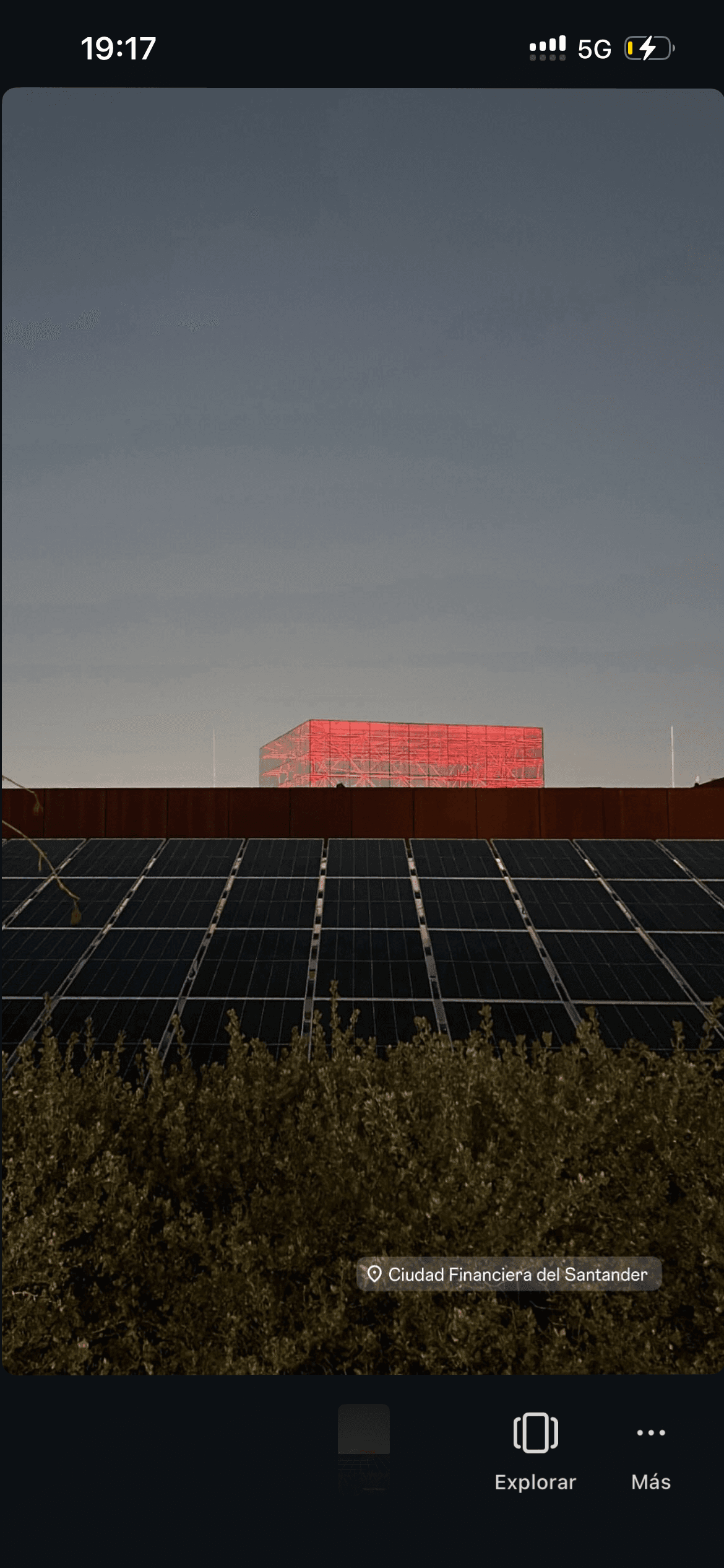 Paneles solares y arbustos frente a un edificio con estructura roja iluminada al atardecer.